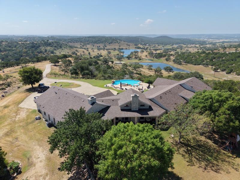 Drone view of a large shingle-roof home with a pool and ponds overlooking a wide Hill Country landscape