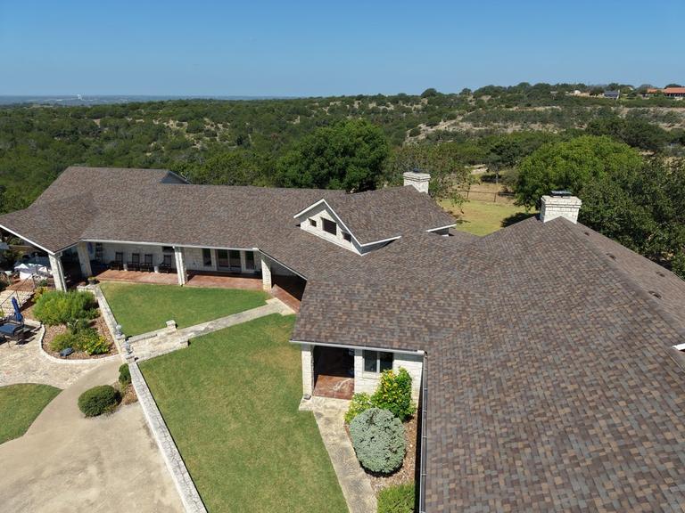 Drone view of a large stone house with a brown multi-tone architectural shingle roof, green lawn, and a Texas Hill Country view